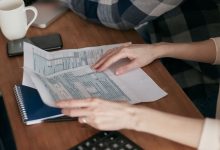 Mãos organizam formulários fiscais sobre uma mesa ao lado de uma calculadora, computador e caderno.