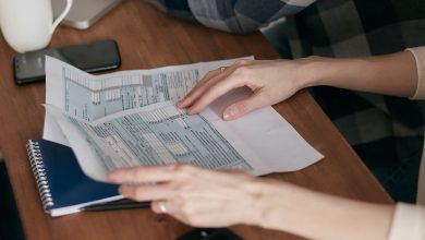 Mãos organizam formulários fiscais sobre uma mesa ao lado de uma calculadora, computador e caderno.