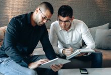 Dois profissionais analisam informações em um tablet e fazem anotações em um caderno durante uma reunião de trabalho.