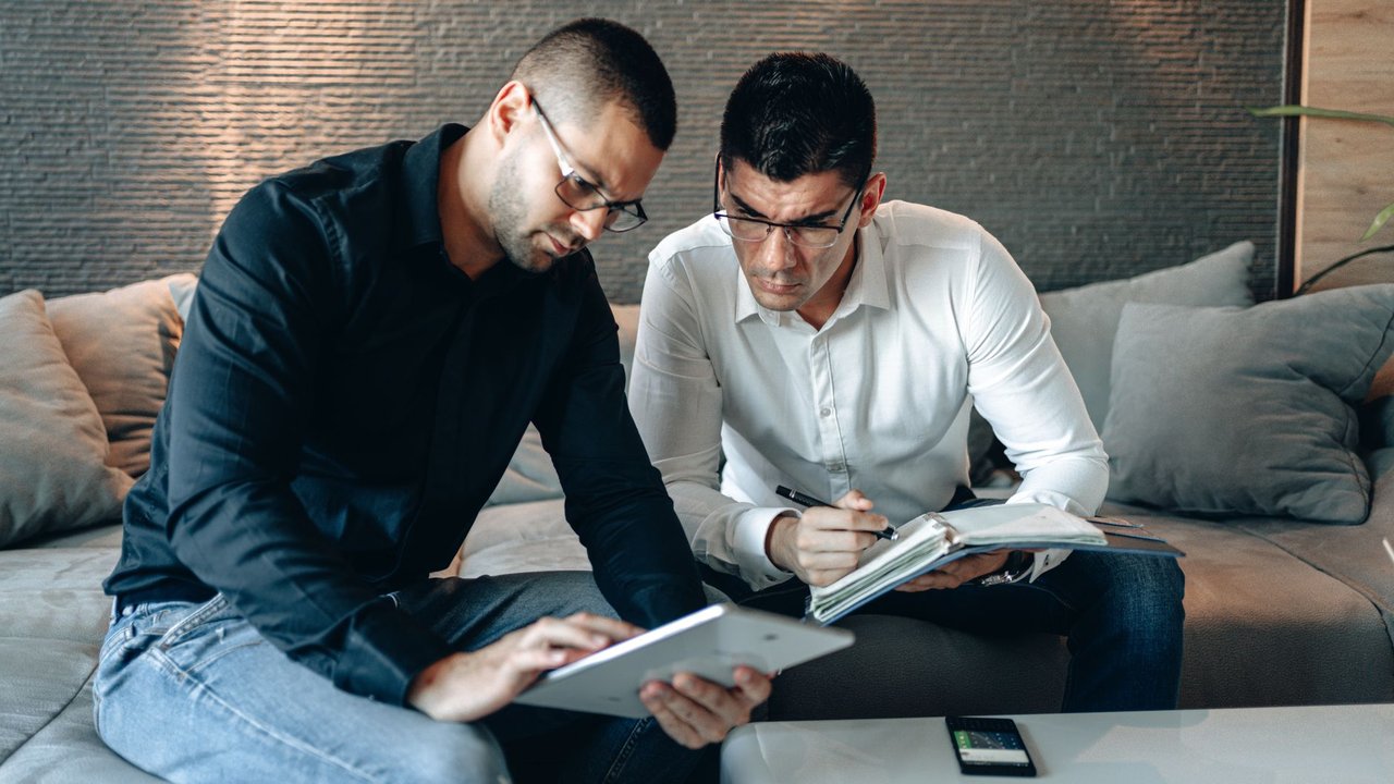 Dois profissionais analisam informações em um tablet e fazem anotações em um caderno durante uma reunião de trabalho.