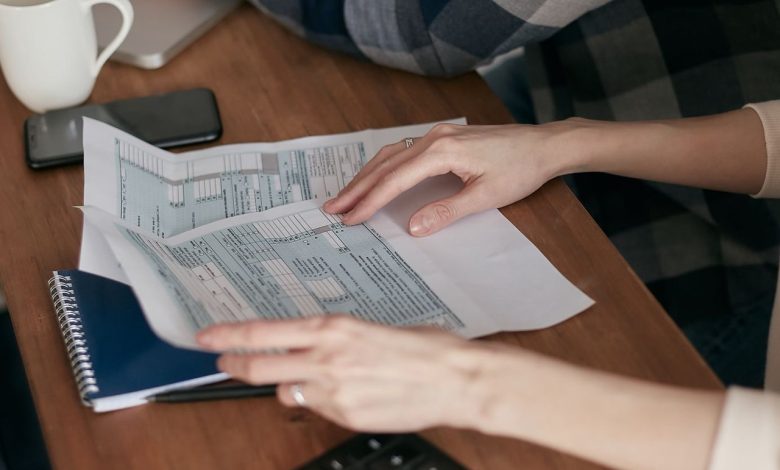 Mãos organizam formulários fiscais sobre uma mesa ao lado de uma calculadora, computador e caderno.