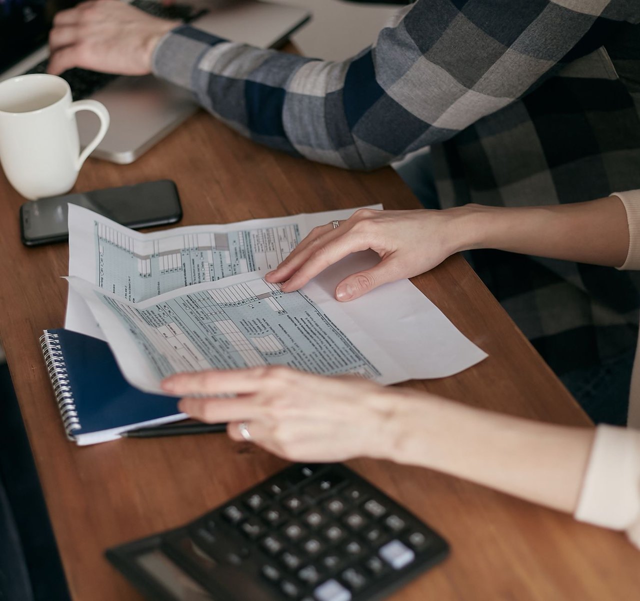Mãos organizam formulários fiscais sobre uma mesa ao lado de uma calculadora, computador e caderno.